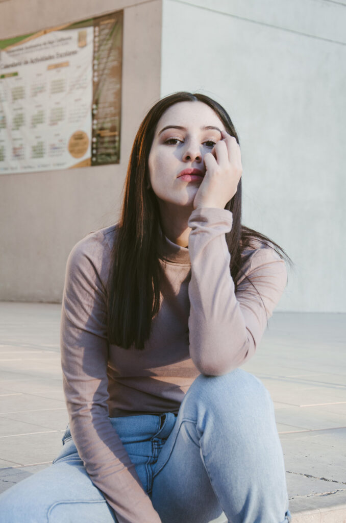 Gabriela Inzunza sits outdoors posing with her hand on her face wearing a beige turtleneck and blue jeans.